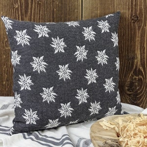 Puede incluir: Un cojín cuadrado con un fondo gris oscuro y un patrón floral blanco repetido. El cojín está sobre una tela a rayas blancas y grises. El fondo es una pared de madera.