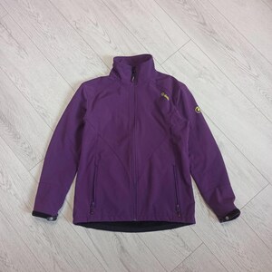 May include: A purple softshell jacket featuring a high collar, a full-length front zip, and two zipped side pockets. The jacket includes a small yellow logo on the upper chest and left sleeve, with black adjustable cuffs and a black hem.