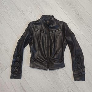 May include: A black leather jacket featuring a stand-up collar, a central front zipper, and two zippered chest pockets. Each sleeve is detailed with three horizontal straps, each secured with a small metal buckle. The interior label reads Gucci.