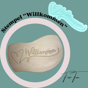 Könnte beinhalten: Ein hellblauer Ausstecher und ein gestempelter Abdruck des Wortes "Willkommen" mit einem Herzdesign. Der Text "Stempel 'Willkommen'" befindet sich über dem Abdruck, und die Signatur "Für Jon" darunter.