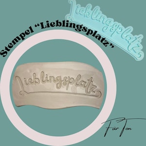 Op de afbeelding: Een lichtblauwe stempel en een klei-afdruk, beide met het woord "Lieblingsplatz" in een cursief lettertype. De stempel staat rechtsboven en de afdruk in het midden. De achtergrond is teal.