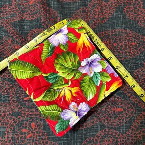 Puede incluir: Un salvamanteles cuadrado rojo acolchado con un estampado floral tropical. El diseño presenta hojas verdes, hibiscos morados y flores amarillas y naranjas. El salvamanteles mide aproximadamente 15 cm por 15 cm.