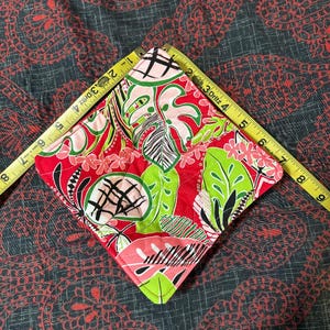 May include: A square, quilted fabric coaster with a vibrant tropical floral pattern in red, green, pink, and black. The coaster measures approximately 4 inches across. A yellow measuring tape is placed diagonally across the coaster.