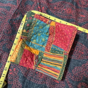 Puede incluir: Un cuadrado acolchado de patchwork con telas rojas, azules, verdes y amarillas. El cuadrado mide aproximadamente 23 cm de ancho, medido con una cinta métrica amarilla. El fondo es una tela oscura con un estampado floral rojo.