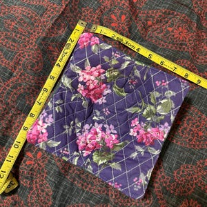 May include: A square, quilted pot holder with a floral pattern in shades of pink and purple. The pot holder is approximately 7 inches square. A yellow measuring tape is placed diagonally across the pot holder.