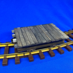 May include: A wooden platform train carriage on a brown and gold model railway track. The carriage is made of wooden planks and has a brown metal frame.