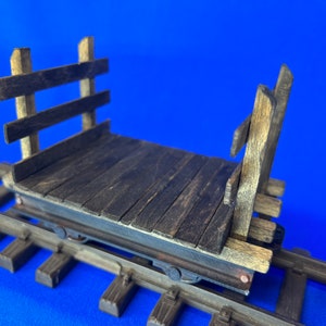May include: A wooden toy train carriage with a flatbed and wooden sides. The carriage is on a set of train tracks.