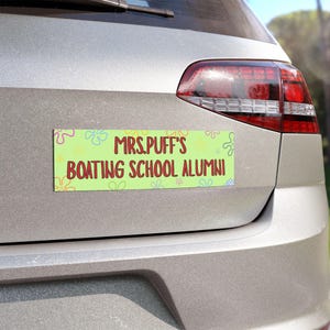 Może przedstawiać: Prostokątny magnes na samochód z napisem "MRS. PUFF'S BOATING SCHOOL ALUMNI" w czerwonych literach na jasnozielonym tle. Magnes jest ozdobiony kwiatami z kreskówek i jest przymocowany do tyłu srebrnego samochodu.