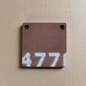 以下が含まれることがあります： 上部に2つの丸い穴がある、正方形の赤茶色の金属板。番号「477」は、プレートの下部に白でステンシルされています。プレートは、明るい茶色の段ボールの背景に配置されています。
