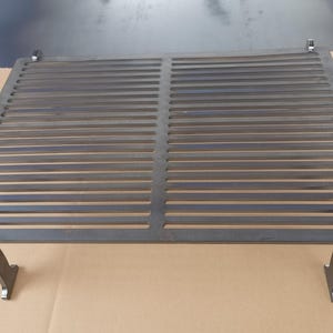 May include: A metal grill grate with a rectangular shape and a set of two metal legs. The grate has a black finish and is made of metal.