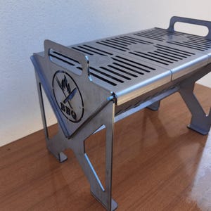 Pode incluir: Uma pequena grelha para churrasco prateada com uma superfície de cozedura ranhurada e pegas integradas. A grelha apresenta um logótipo com uma chama e uma faca, e a palavra "BBQ". As pernas são anguladas para estabilidade. Esta grelha portátil foi concebida para cozinhar ao ar livre.