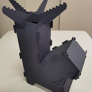 May include: A dark grey metal camping stove with a rectangular base and a chimney-like structure. The top has four angled supports with notched edges. The stove is designed for outdoor cooking and camping.