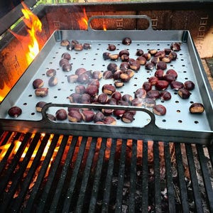 May include: A metal roasting pan with holes sits on a grill over an open fire. The pan is filled with chestnuts roasting over the flames.