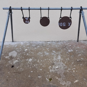 May include: A metal shooting target stand with three circular targets hanging from a horizontal bar. The targets are brown with white numbers and letters. The stand is made of gray metal and has a tripod base.