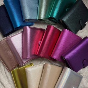 Könnte beinhalten: Eine Sammlung farbenfroher, metallisch glänzender Personal Organizer. Die Organizer sind in verschiedenen Farbtönen erhältlich, darunter Blau, Grün, Rosa, Lila, Gold und Silber. Jeder Organizer hat einen Schnappverschluss.