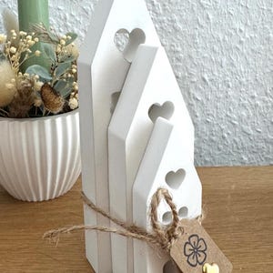 Könnte beinhalten: Ein Stapel aus drei weißen Holzhausdekorationen mit herzförmigen Ausschnitten. Die Häuser sind mit Bindfaden zusammengebunden und haben ein kleines Etikett mit einem Blumen- und Herzdesign. Eine dekorative Kerze und ein Blumenarrangement befinden sich im Hintergrund.