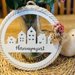 Könnte beinhalten: Weißer Wandbehang aus Holz mit einem Seilrand, der ein ausgeschnittenes Stadtbilddesign und den Text "Hereinspaziert" zeigt. Eine kleine Vogelfigur und eine Vase mit Blumen sind ebenfalls vorhanden.