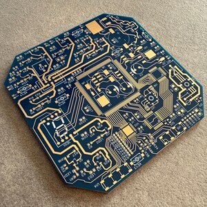 May include: A blue and gold circuit board featuring detailed electronic pathways and components. The board has a distinctive, geometric shape and is designed with a complex network of lines and symbols, showcasing a technological theme.