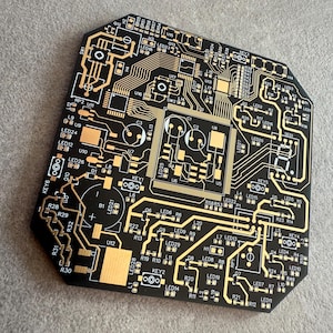 May include: Black circuit board with gold electrical pathways and components. The board features various labelled components, including LEDs, resistors, and integrated circuits. The design is intricate and geometric, with a modern aesthetic.