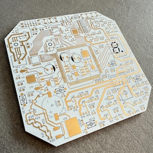 May include: A white printed circuit board with detailed gold-coloured circuitry. The board includes various components, such as LED indicators, and labelled sections. The design is complex, with numerous lines and shapes, suggesting a technological or electronic device.