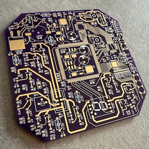 May include: A purple circuit board with gold-coloured traces and components. The board features various electronic components, including LEDs, resistors, and integrated circuits. The design is intricate, with numerous pathways and connection points, showcasing a complex electronic system.