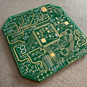 May include: A green printed circuit board featuring gold-coloured circuitry and components. The board's intricate design includes integrated circuits, resistors, and capacitors. The square board has rounded corners and is a piece of electronic hardware.