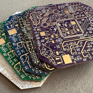 May include: A stack of colorful circuit boards with intricate gold-colored traces and components. The boards are in shades of white, green, blue, and purple. The boards have a unique octagonal shape.