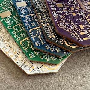 May include: A collection of circuit boards in various colours, including green, blue, and purple, with gold-coloured circuitry. The boards are stacked, revealing their angled edges. The bottom board is beige, and the boards are covered in small components.