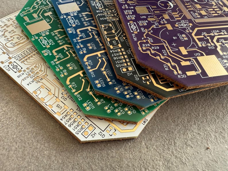 May include: A collection of circuit boards in various colours, including green, blue, and purple, with gold-coloured circuitry. The boards are stacked, revealing their angled edges. The bottom board is beige, and the boards are covered in small components.