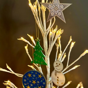 May include: A white twig Christmas tree with lights, decorated with four ornaments: a green Christmas tree, a blue and gold star ornament, a gold snowman ornament, and a gold star ornament.