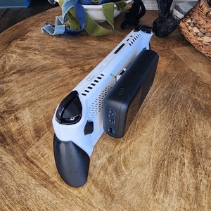 Op de afbeelding: Een wit en zwart handheld gaming-apparaat met een zwarte powerbank bevestigd. Het apparaat heeft een getextureerde grip en een scherm. De powerbank heeft meerdere USB-poorten. De items staan op een houten tafel.