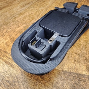 May include: A black 3D printed case with a compartment for a power adapter and a cord. The case has a textured surface and a black cord is wrapped around it.