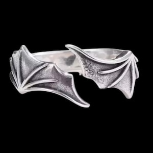 Może przedstawiać: Srebrny pierścionek z motywem skrzydeł nietoperza. Pierścionek posiada dwa szczegółowe skrzydła, które rozciągają się na zewnątrz, tworząc unikalną i gotycką estetykę. Pierścionek jest umieszczony na czarnym tle.