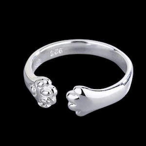 May include: A silver-colored ring with a paw design. The ring features a detailed paw print on one end and a smooth paw shape on the other, creating an open design. The ring is set against a black background.
