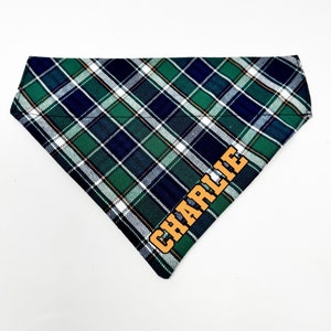 May include: A green, blue, and white plaid dog bandana with the name "CHARLIE" embroidered in orange on the bottom right corner.