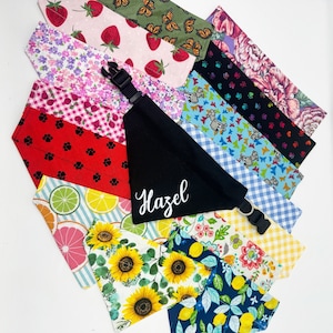 May include: A collection of colorful dog bandanas with various patterns, including floral, geometric, and animal prints. One bandana is black with the name "Hazel" embroidered in white.