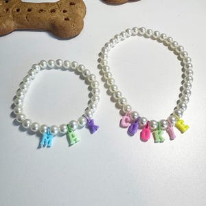 May include: Two pearl necklaces with colourful letter charms spelling out the names "Max" and "Cookie".