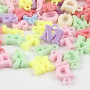 May include: Assortment of colourful plastic letter charms in pastel shades of pink, yellow, green, and purple. The letters are all uppercase and have a hole for stringing.