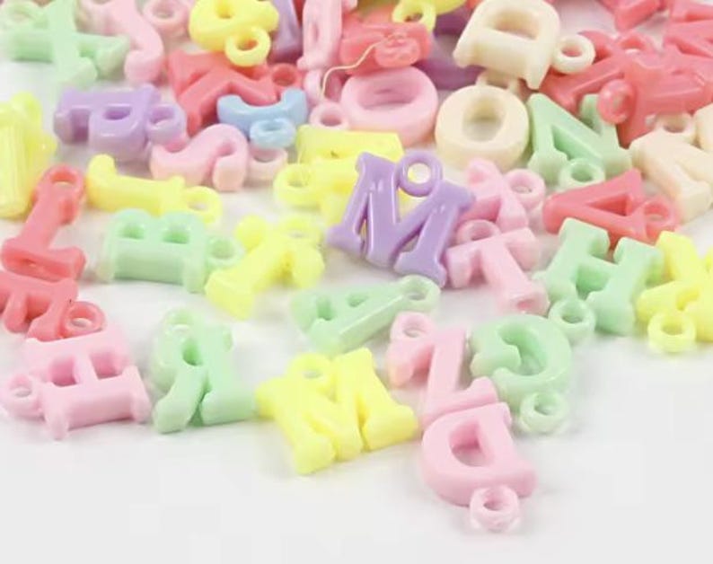 May include: Assortment of colourful plastic letter charms in pastel shades of pink, yellow, green, and purple. The letters are all uppercase and have a hole for stringing.