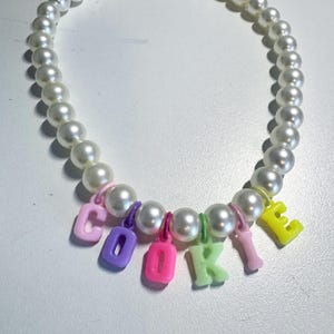 May include: A white pearl necklace with colourful letter charms spelling out the word "COOKIE".