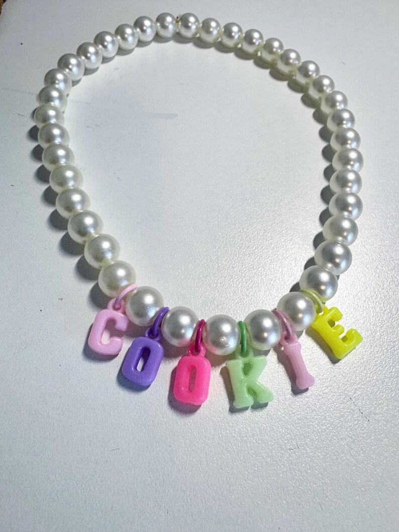 May include: A white pearl necklace with colourful letter charms spelling out the word "COOKIE".