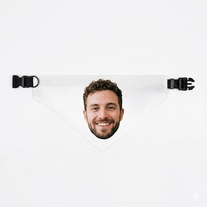 Op de afbeelding: Witte driehoekige bandana voor huisdieren met een afdruk van het gezicht van een lachende man. De bandana heeft zwarte plastic verstelbare gespen aan elk uiteinde. De achtergrond is effen wit.