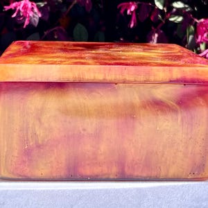 May include: A square, wooden box with a lid. The box is made of resin and has a marbled, swirled pattern in shades of orange, yellow, and red.