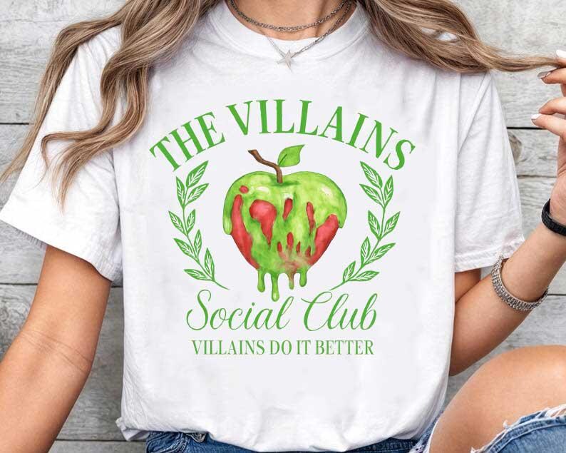 May include: White t-shirt with a green and red apple graphic. The text on the shirt reads "THE VILLAINS SOCIAL CLUB" and "VILLAINS DO IT BETTER" in green. The apple design is surrounded by green leaves.