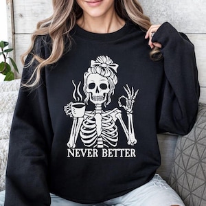 K&ouml;nnte beinhalten: Schwarzer Pullover mit einem wei&szlig;en Grafikdruck eines Skeletts, das eine Kaffeetasse h&auml;lt und "Never Better" sagt.
