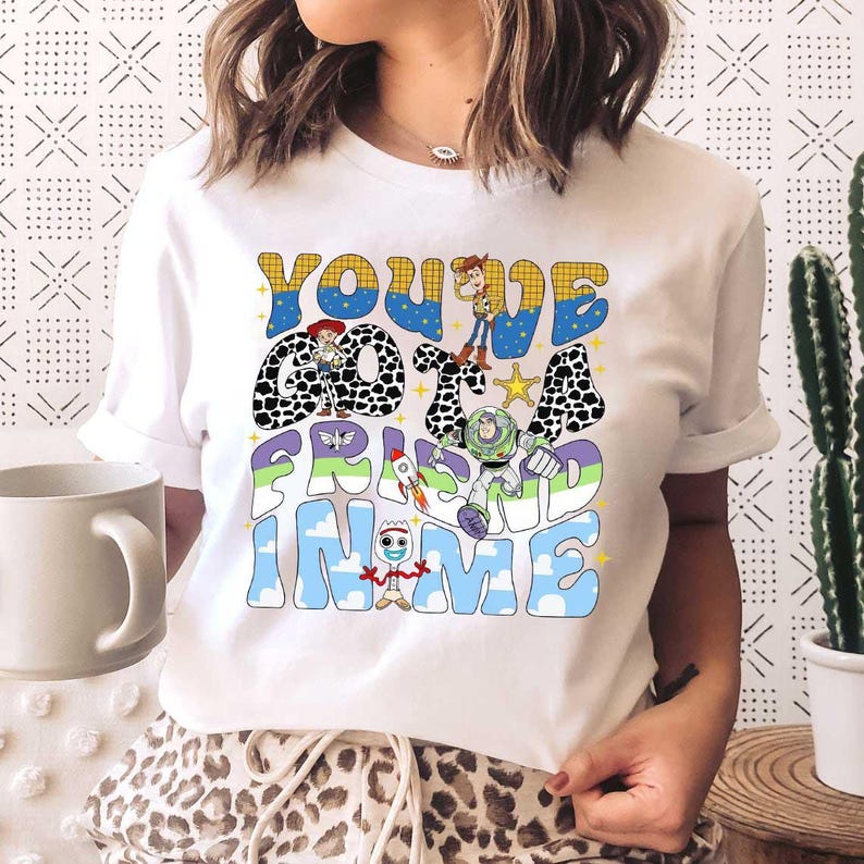以下が含まれることがあります： レトロなフォントで「You've Got a Friend in Me」というフレーズがプリントされた白いTシャツ。デザインには漫画のキャラクターとロケットが含まれています。袖は折り返されています。