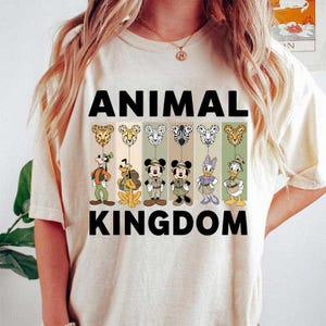 May include: Cream-colored t-shirt with the words "ANIMAL KINGDOM" in black block letters. The shirt features cartoon characters and animal-themed balloons. The balloons have animal print designs, and the characters are in various poses.
