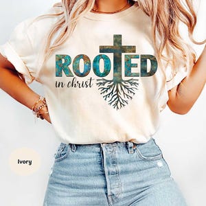 Op de afbeelding: Ivoor kleurig T-shirt met de tekst "ROOTED in christ" in een blauwgroen en bruin ontwerp. Het ontwerp bevat een kruis en een wortelstelsel. Het shirt wordt gecombineerd met een blauwe jeans.