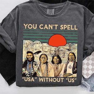 Könnte beinhalten: Dunkelgraues T-Shirt mit dem Text "YOU CAN'T SPELL USA WITHOUT US". Das Shirt zeigt eine Illustration von indianischen Figuren und Mount Rushmore mit einer roten Sonne im Hintergrund. Das Shirt hat einen Rundhalsausschnitt.
