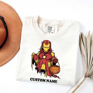 May include: Off-white t-shirt with a Halloween Iron Man design. The graphic depicts Iron Man in a costume holding a jack-o'-lantern bucket. The text "CUSTOM NAME" is printed at the bottom. The shirt is from Comfort Colors.